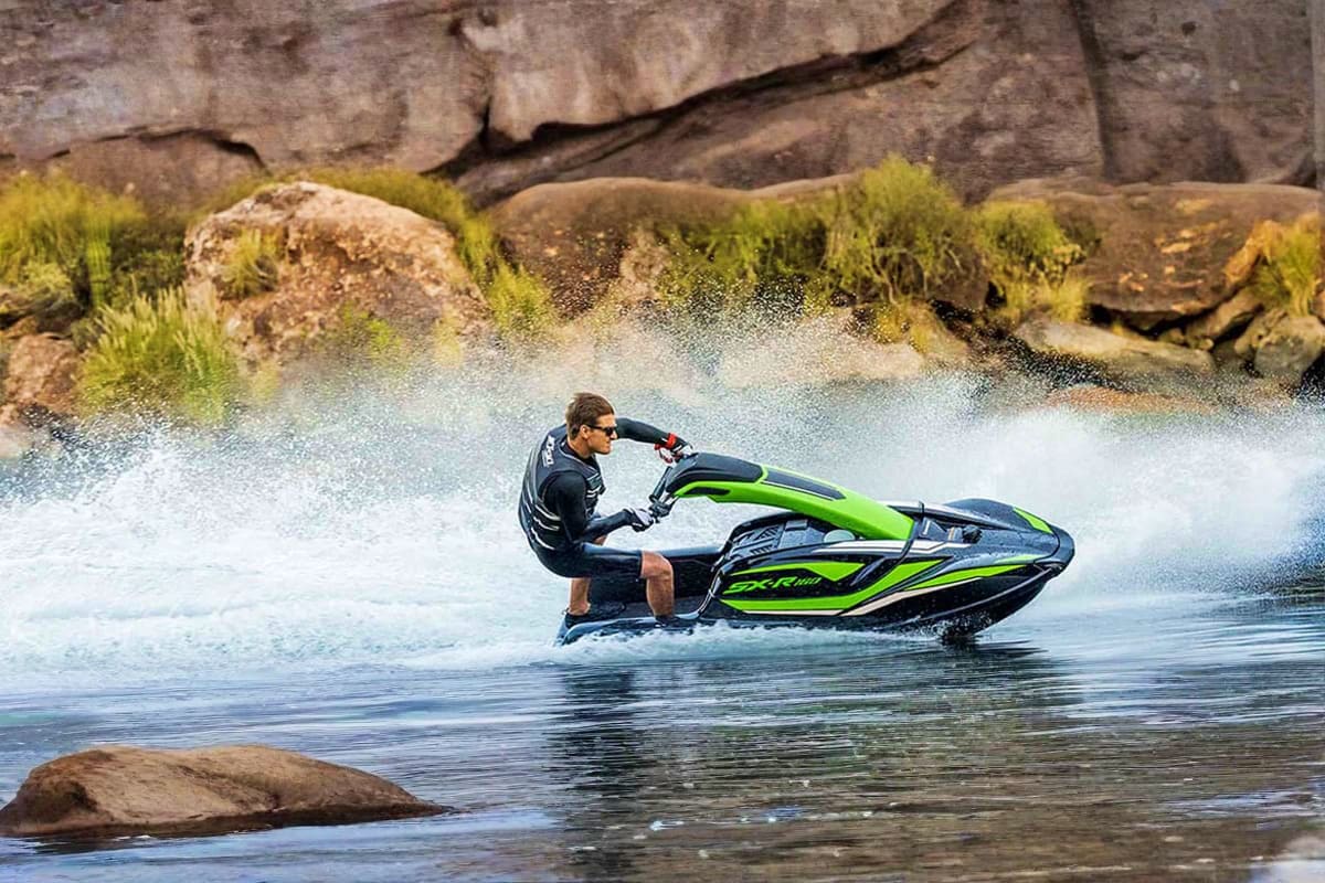 Man riding a green and black jet ski in shallow river water with moss-covered rocks and cliff formations in the background
