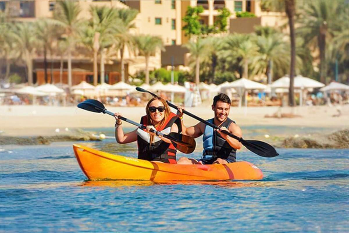 Kayaking Experience in Dubai Couple kayaking in bright orange kayak on blue water with Dubai beach, palm trees, and beachfront buildings in background