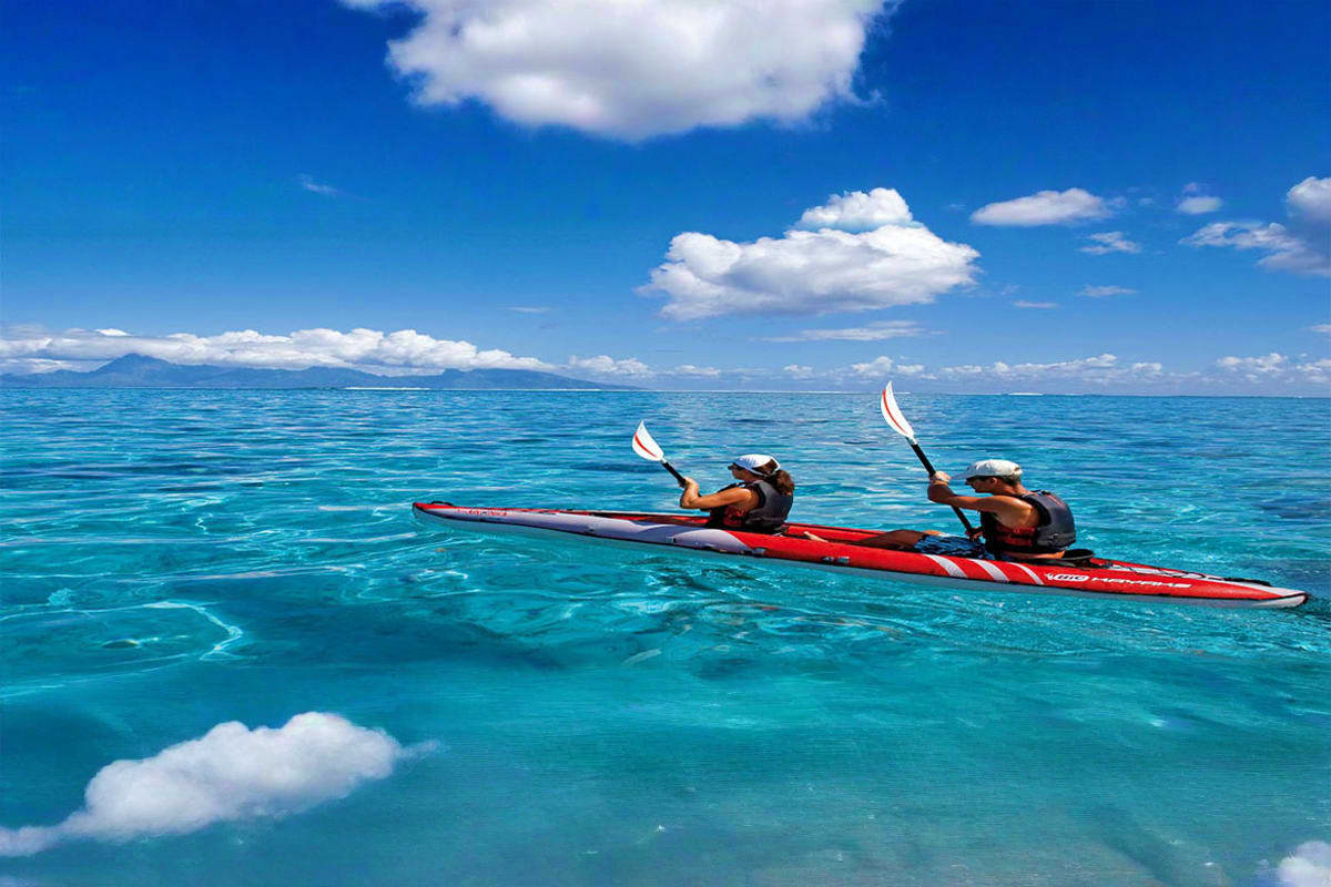 Kayaking Experience in Dubai Two people kayaking in turquoise ocean water with paddles raised, mountains visible on horizon under blue sky with white clou