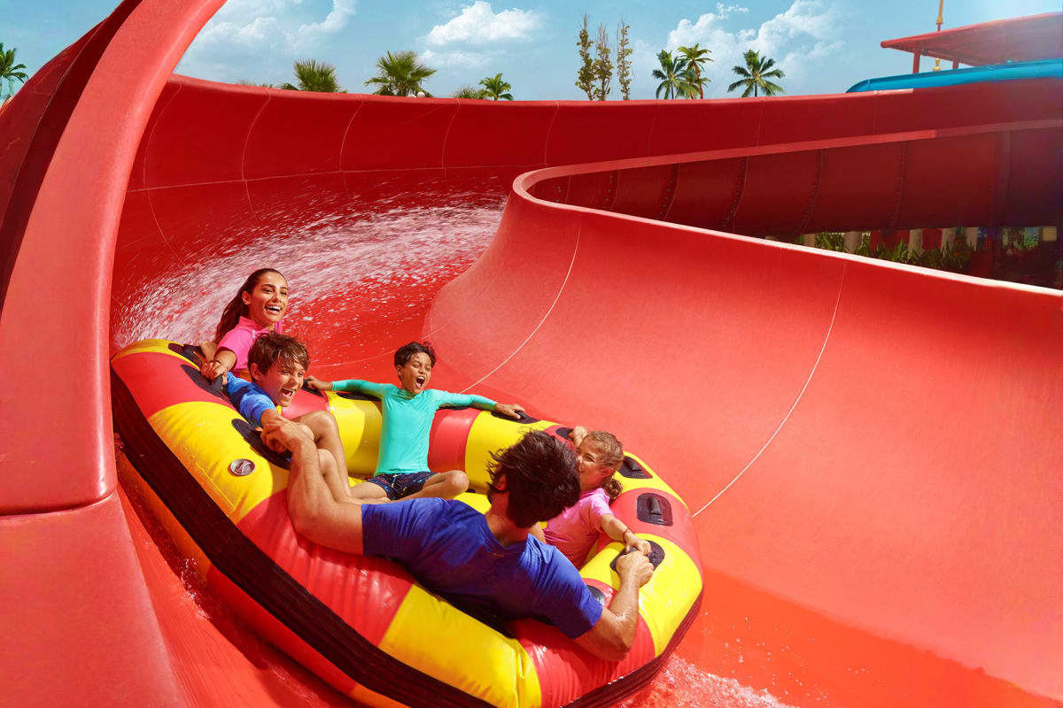 Family riding down red water slide on yellow inflatable raft at water park with palm trees in background