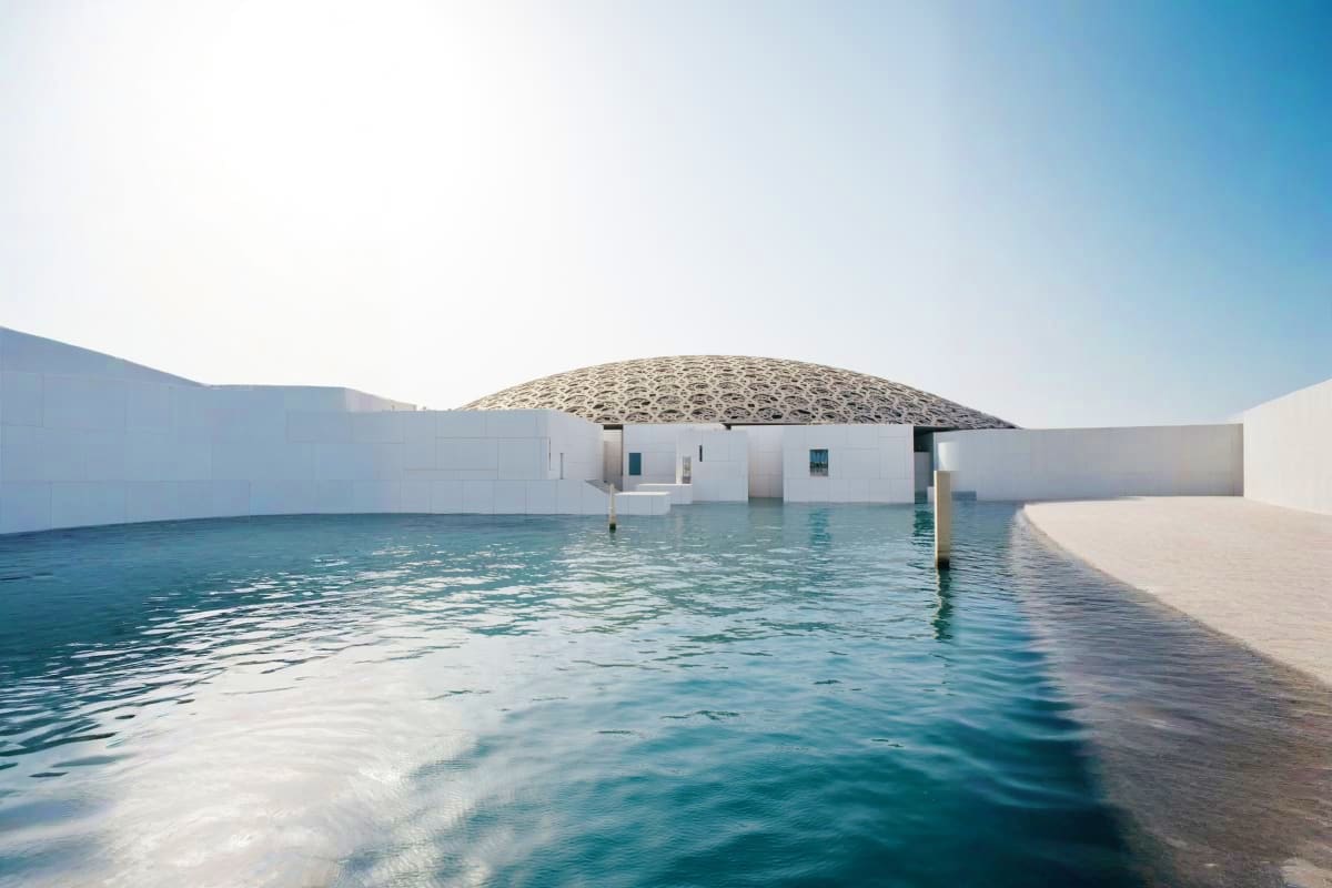 Modern white building with distinctive dome-shaped roof reflected in turquoise water at Louvre Abu Dhabi museum