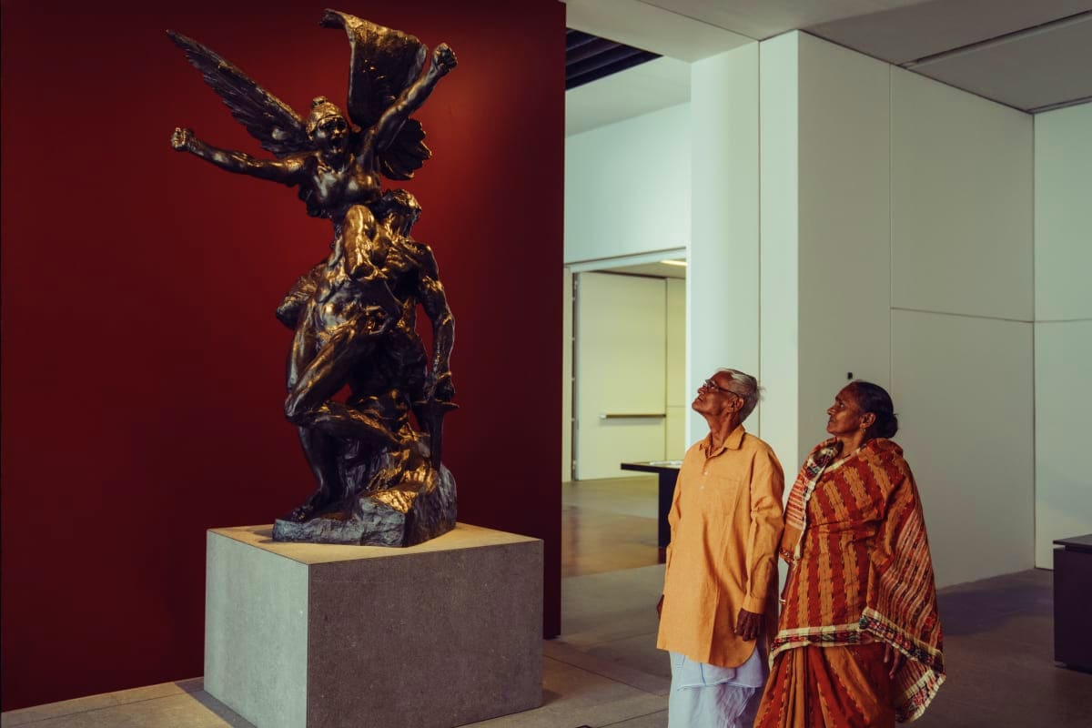 Two visitors viewing a bronze sculptural artwork depicting a winged figure at the Louvre Abu Dhabi museum