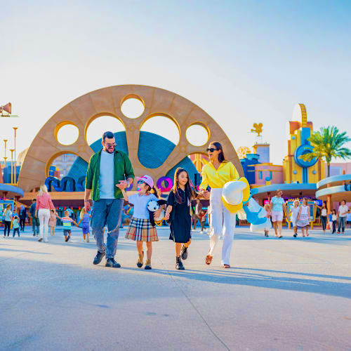 Family walking through Motiongate Dubai theme park entrance with large paw print archway and colorful attractions in backgrou