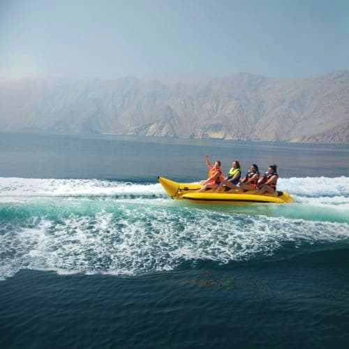 Four tourists riding yellow banana boat in ocean waves with dramatic cliff mountains in background at Musandam Dibba