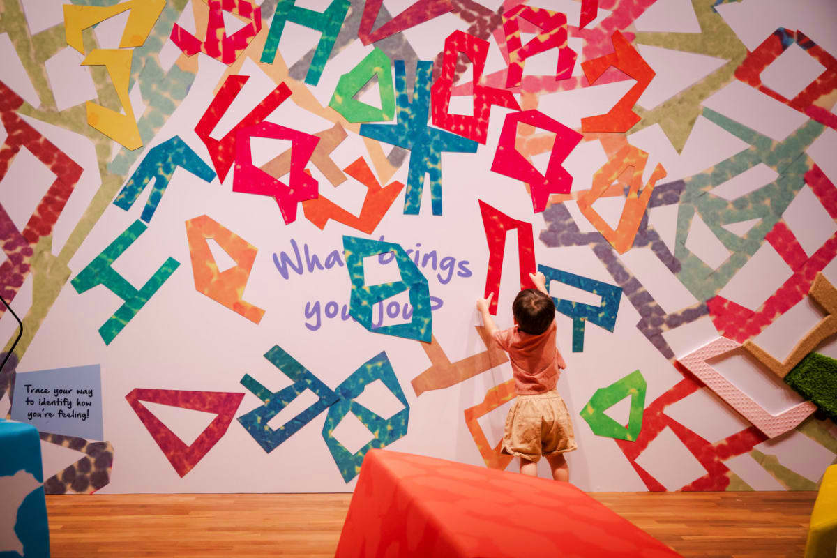 Child interacting with colorful geometric art installation displaying "What brings you joy?" at National Gallery Singapore