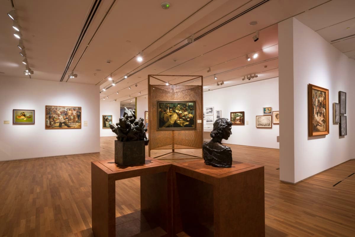 Modern art gallery interior with classical sculptures on pedestals, framed paintings on white walls, wooden flooring, and pro