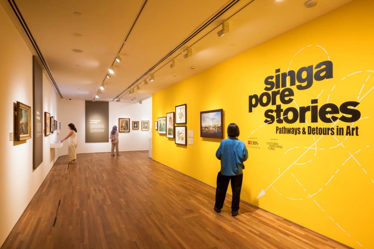 Visitors viewing artworks in a modern gallery space with bright yellow wall displaying "Singapore Stories: Pathways & Detours