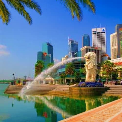 Merlion statue spouting water into lagoon with Singapore skyline skyscrapers, palm trees, and waterfront promenade