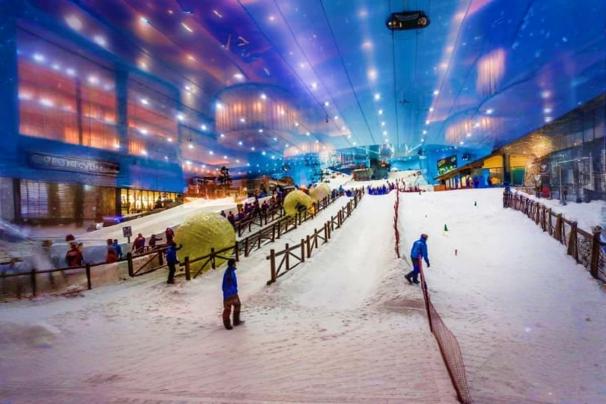 Ski Dubai Tickets Indoor ski slope at Ski Dubai with skiers on white snow, blue ceiling lighting, and retail stores along the sides