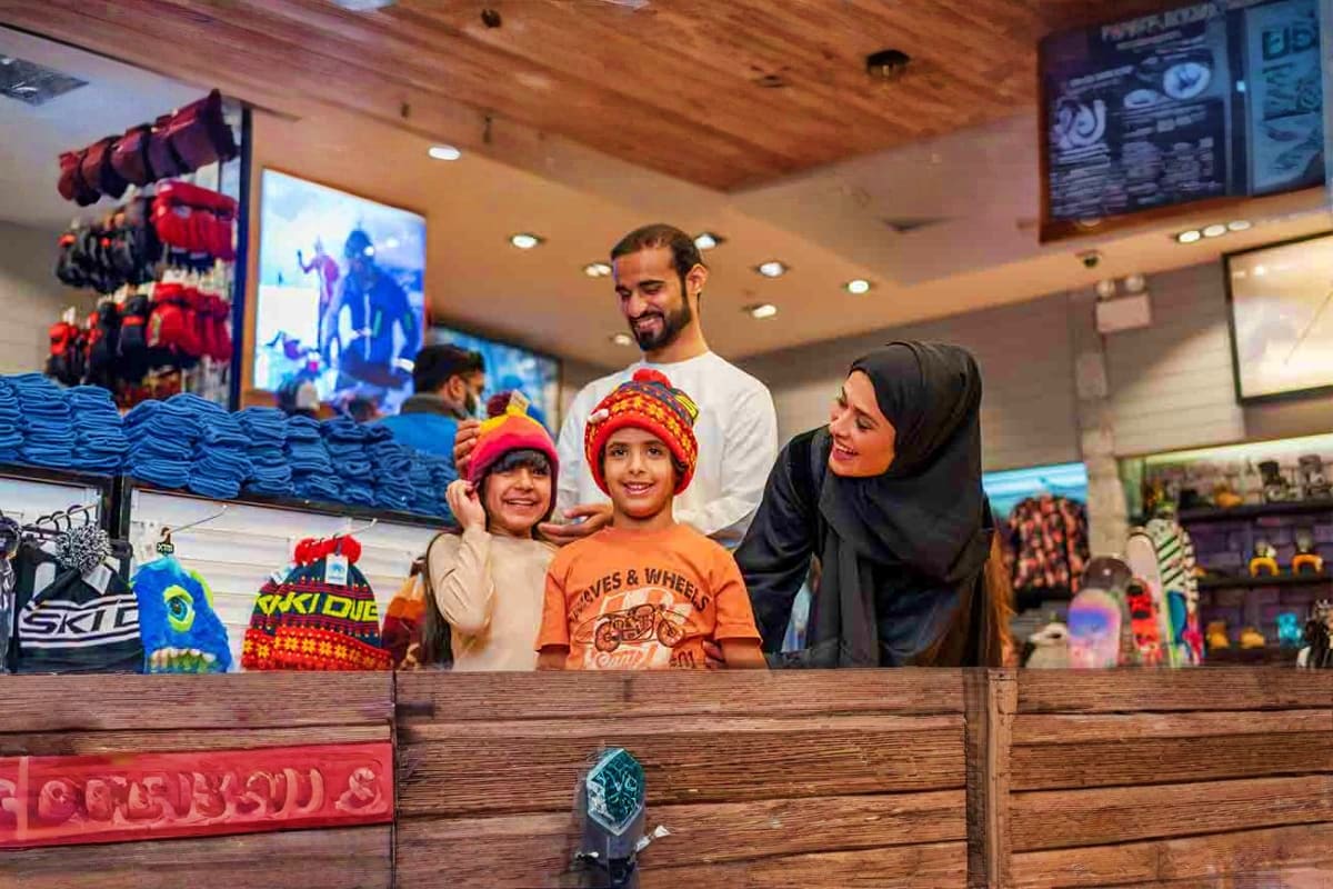 Family trying on colorful winter hats at Ski Dubai indoor ski resort shop counter with snow gear displays