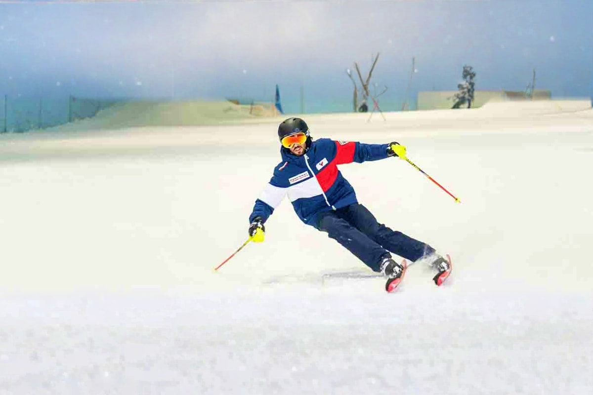 Ski Dubai Tickets Skier in blue and red jacket navigating downhill slope at Ski Dubai indoor snow facility with misty mountains in background