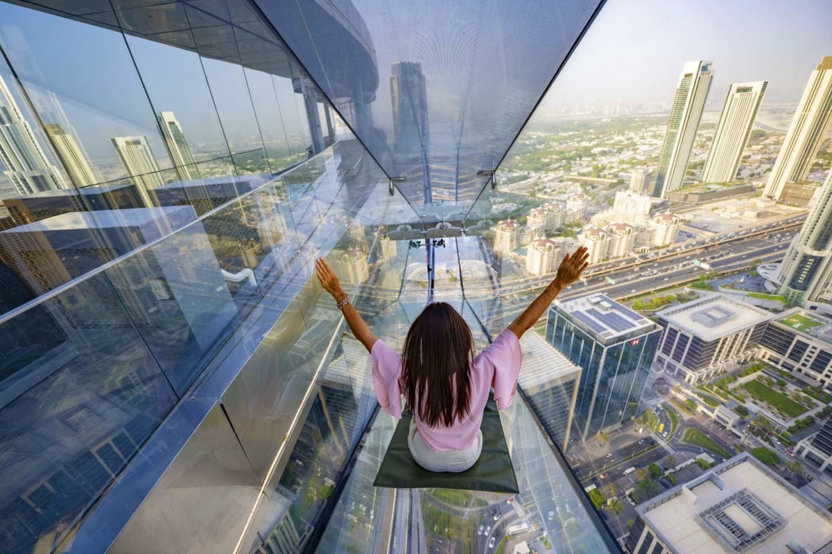 Woman with arms raised sitting in glass floor observation deck overlooking Dubai skyline and cityscape