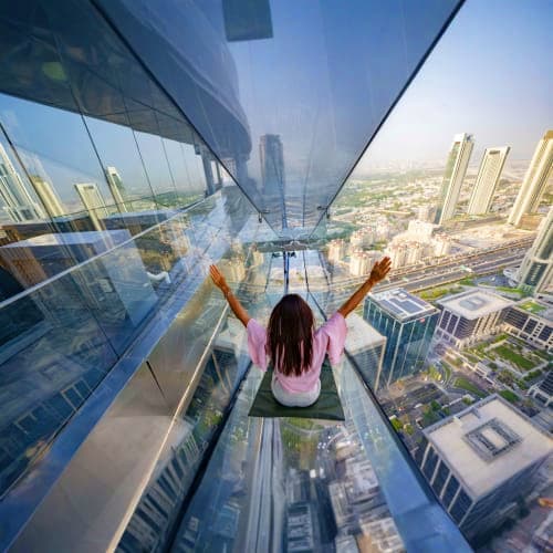Person with arms raised sitting on glass skywalk high above Dubai cityscape and skyscrapers