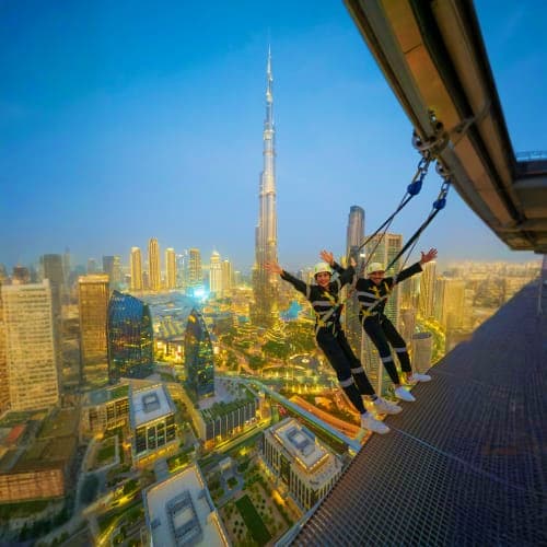 Two people rappelling from high-rise building with Burj Khalifa and Dubai skyline visible in background during daytime