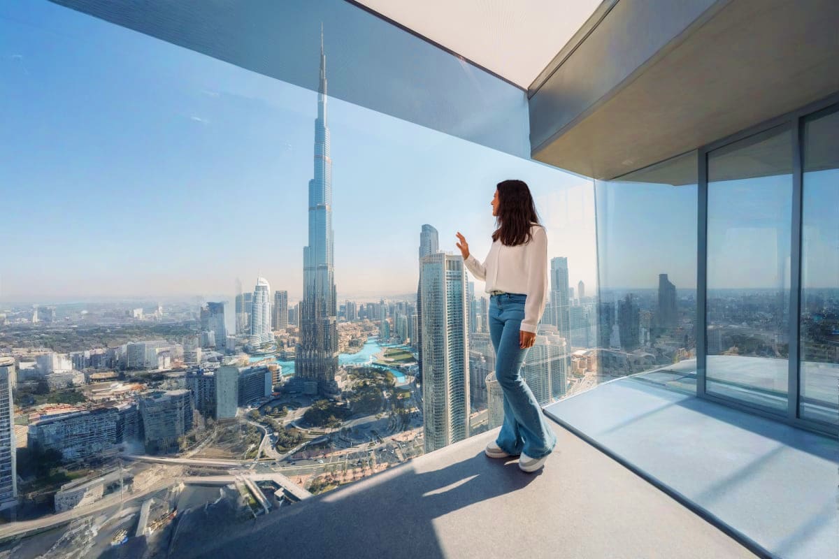 Woman standing on high-rise balcony overlooking Dubai skyline with Burj Khalifa and cityscape visible through floor-to-ceilin