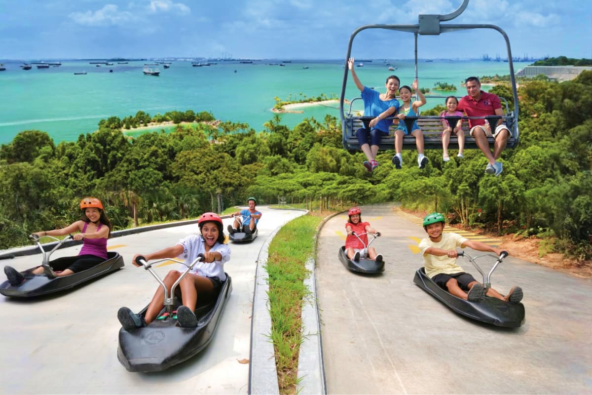 Skyline Luge Singapore Families enjoy luge rides down curved tracks with tropical island and turquoise ocean views in background at Sentosa Island