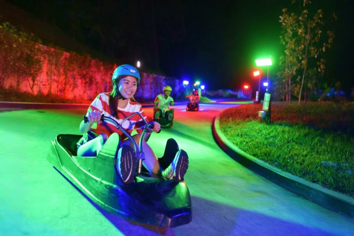 Skyline Luge Singapore Woman riding neon-lit luge down illuminated track at night with other riders and colorful LED lighting along the course at Sk