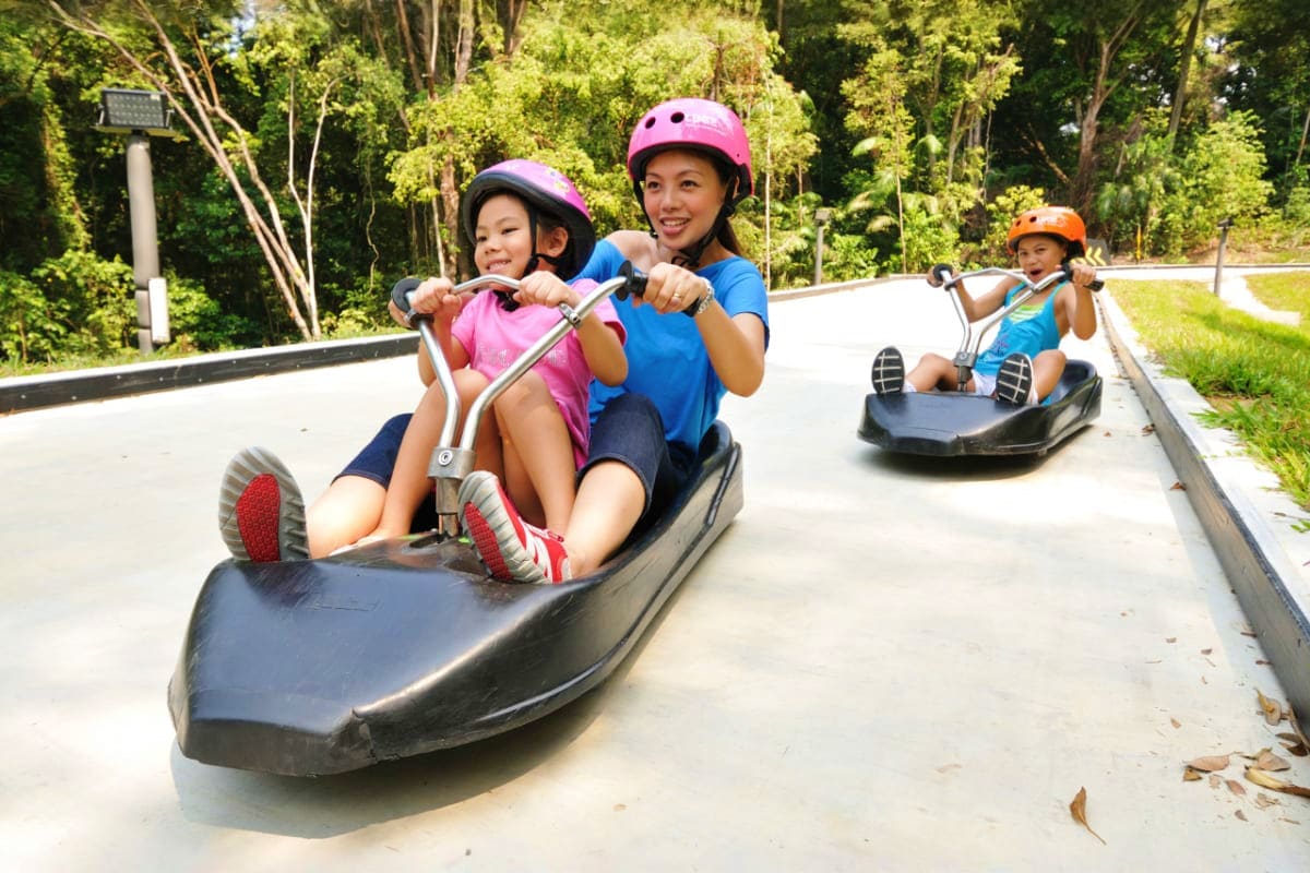 Woman and child riding a luge cart down a concrete track at Skyline Luge Singapore, wearing protective helmets with trees in