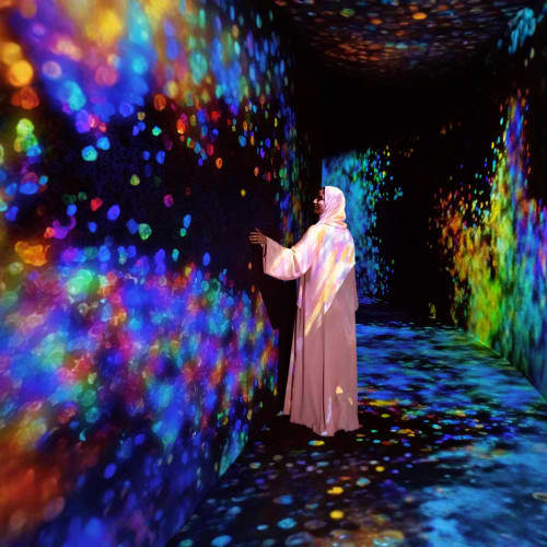 TeamLab Phenomena Person in white robe interacting with immersive digital art installation featuring colorful projected particles and lights on