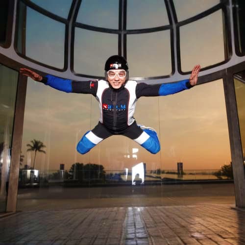 Person indoor skydiving in black and blue suit with arms spread wide inside iFly wind tunnel with Singapore cityscape visible