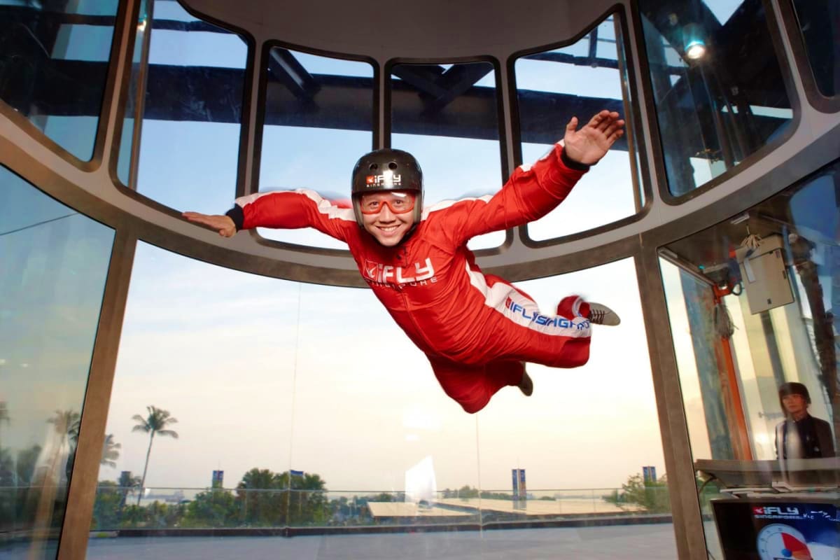 iFly Singapore Child in red flight suit and helmet performing indoor skydiving in wind tunnel at iFly Singapore with city skyline view throu