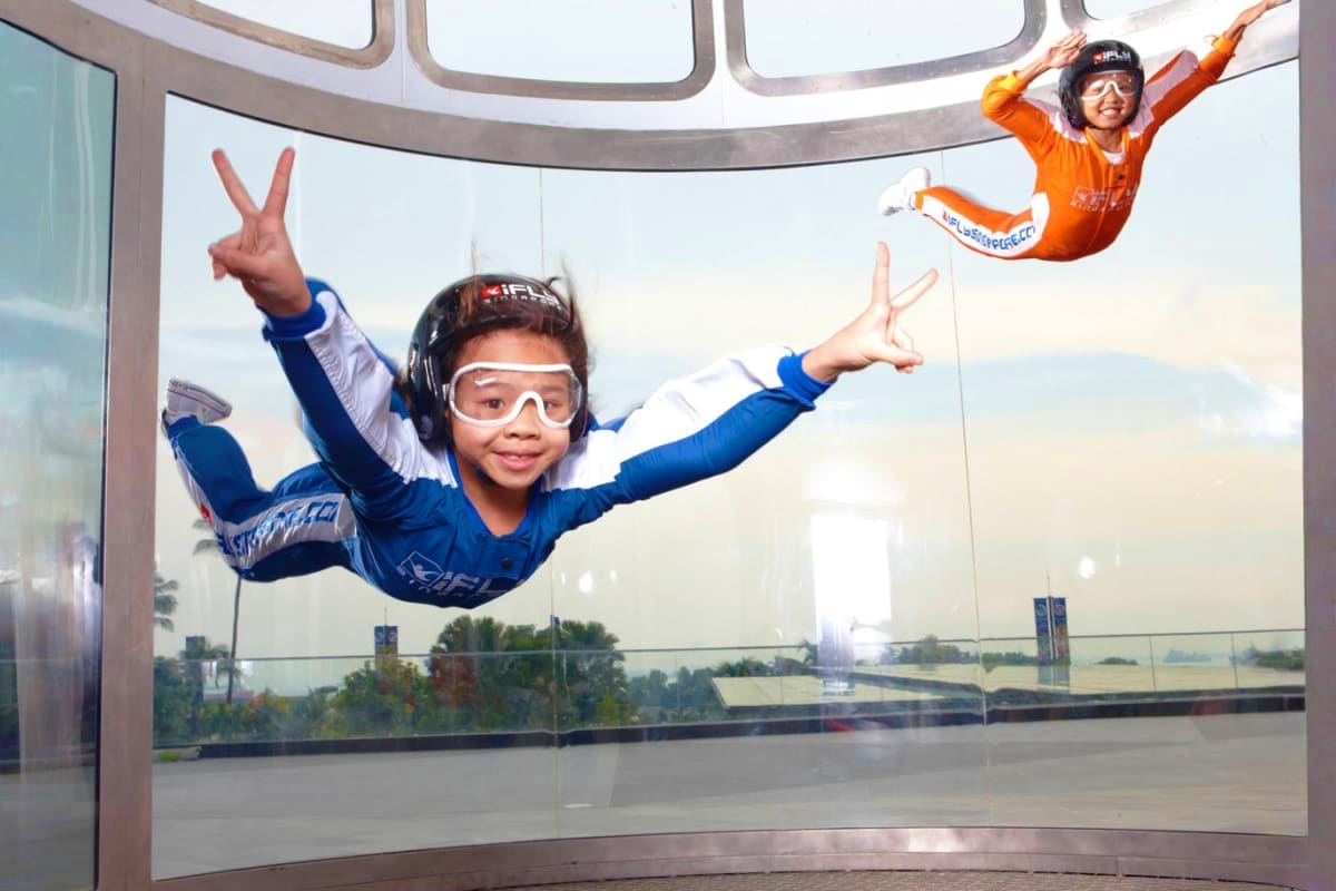 iFly Singapore Two children indoor skydiving at iFly Singapore, one in blue suit making peace sign, another in orange suit floating above, w