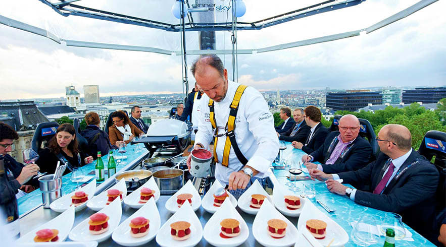 Dinner in the Sky Chef serving plated desserts at suspended dining platform high above London cityscape with guests seated at tables below