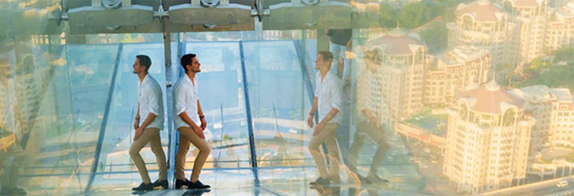 Sky Views Edge Walk Two men standing on transparent glass floor platform high above cityscape with tall buildings visible below and in background