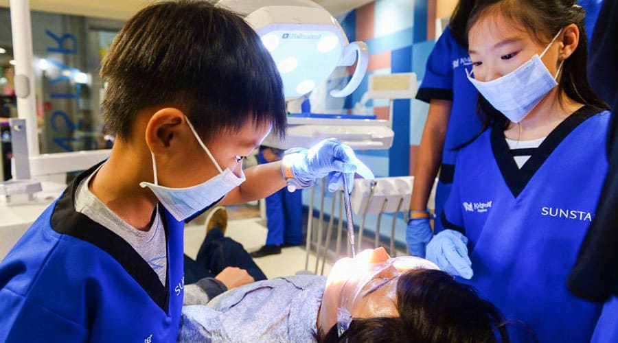 Young visitors wearing blue uniforms and masks practicing dental procedures on a model patient at KidZania Singapore's dentis