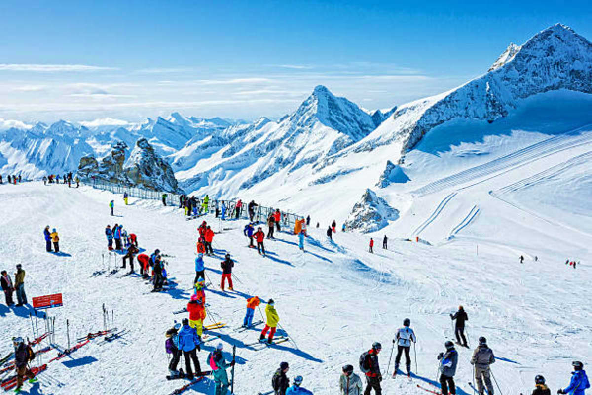 Busy alpine ski resort with snow-covered mountains, skiers on slopes, and clear blue sky in Austria