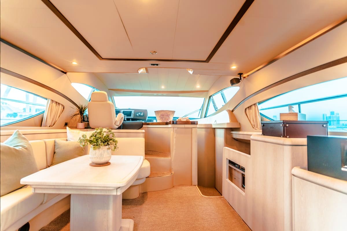 Luxury yacht cabin interior with cream upholstery, dining table, kitchen galley, and panoramic windows overlooking water