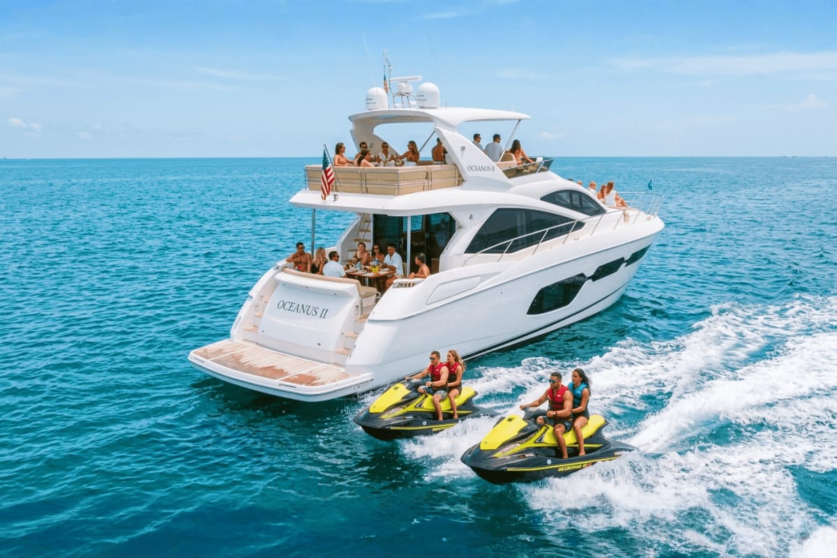 Luxury white yacht with guests on upper and lower decks, two yellow jet skis in foreground on turquoise ocean water with wave