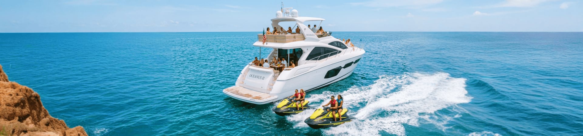 Luxury white yacht with passengers and two yellow jet skis in turquoise waters near rocky coastline