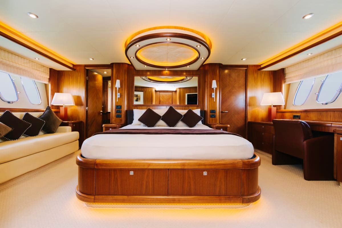 Luxury yacht master bedroom with curved king bed, rich wood paneling, cream seating area, and decorative ceiling lighting