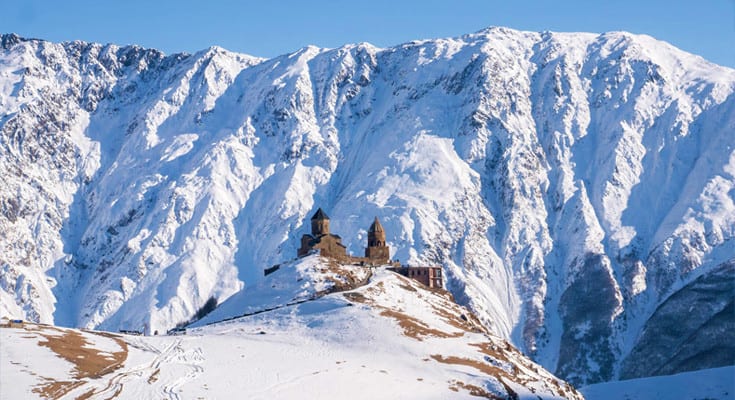 Snow-covered mountain peaks with ancient stone church nestled on hillside in winter landscape, Georgia
