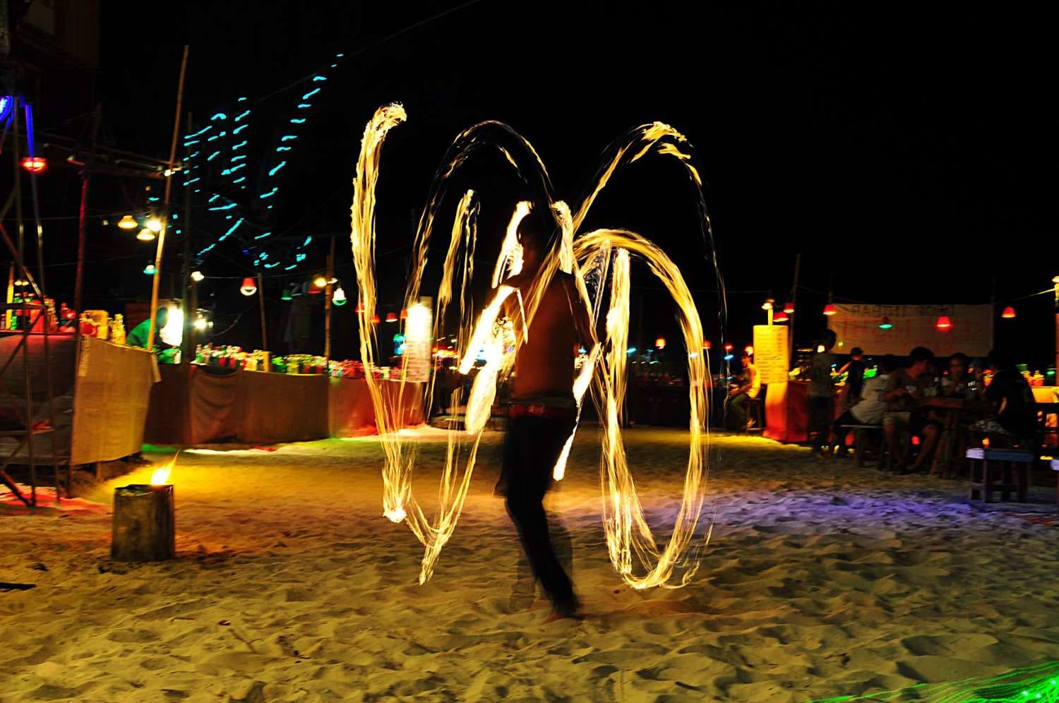 Fire dancer performing with glowing poi at nighttime beach festival with audience seated around sandy arena
