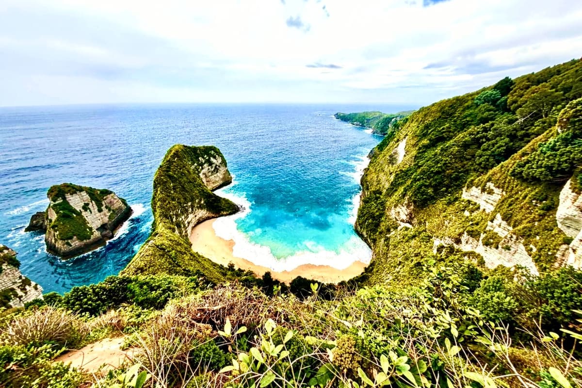 Aerial view of Kelingking Beach in Bali with turquoise waters, white sand cove, and lush green cliffs