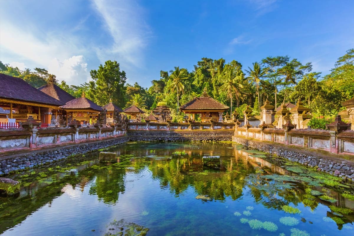Traditional Balinese temple with thatched-roof structures reflected in sacred pool surrounded by lush tropical vegetation