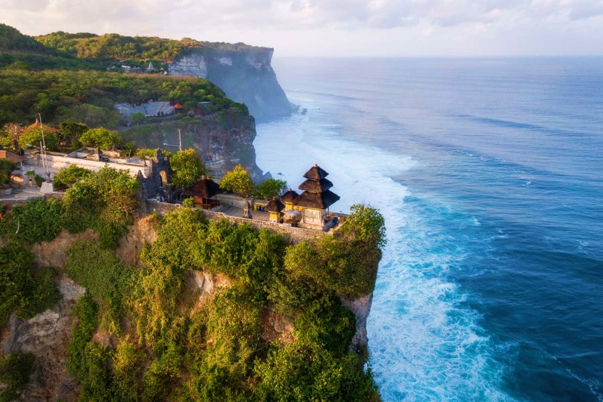 Aerial view of clifftop temple overlooking turquoise ocean waves and lush green vegetation in Bali, Indonesia