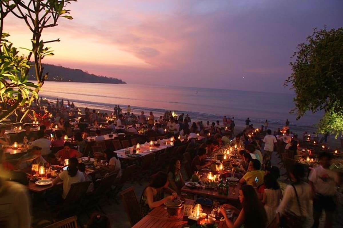 Beachfront dining event at sunset in Bali with candlelit tables, guests, ocean view, and tropical vegetation