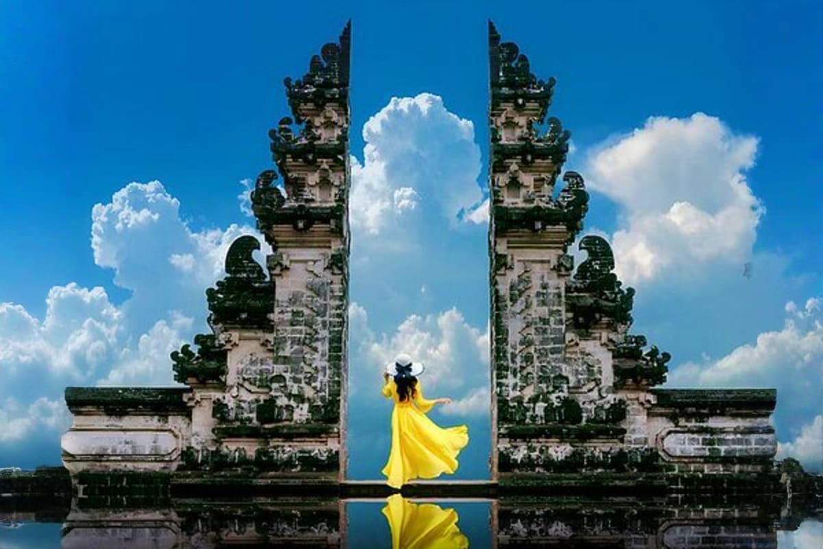Woman in yellow dress posing between ornate Balinese temple gates with reflection in water and blue sky