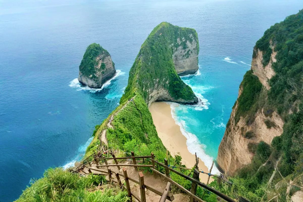 Wooden staircase descending clifftop toward Kelingking Beach with turquoise waters and rock formations in Nusa Tenimur, Bali