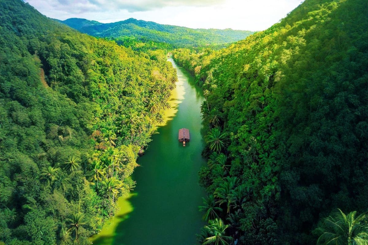 7 Days Manila Clark and Cebu Tour Package Aerial view of a traditional wooden boat on a serene emerald river through dense tropical rainforest and mountains in the Phi
