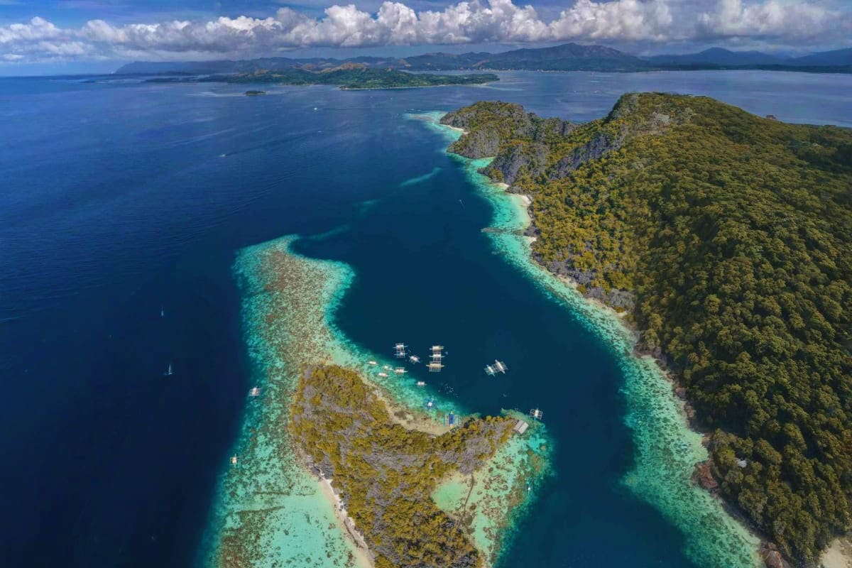 Coron Diving Tour Aerial view of turquoise lagoon surrounded by forested limestone cliffs with anchored boats in Coron, Philippines