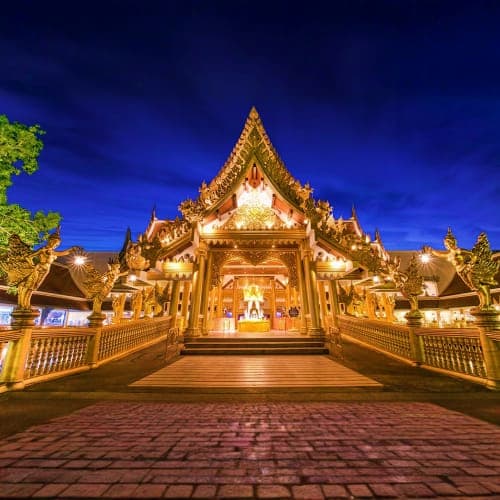 Illuminated golden Thai Buddhist temple with ornate roof and spires at twilight, wooden bridge and courtyard entrance