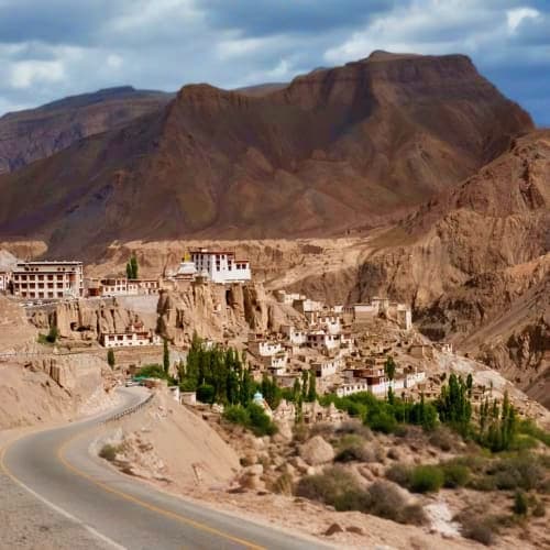 Lamayuru Monastery perched on rocky cliffs overlooking whitewashed village buildings with green poplar trees and winding road