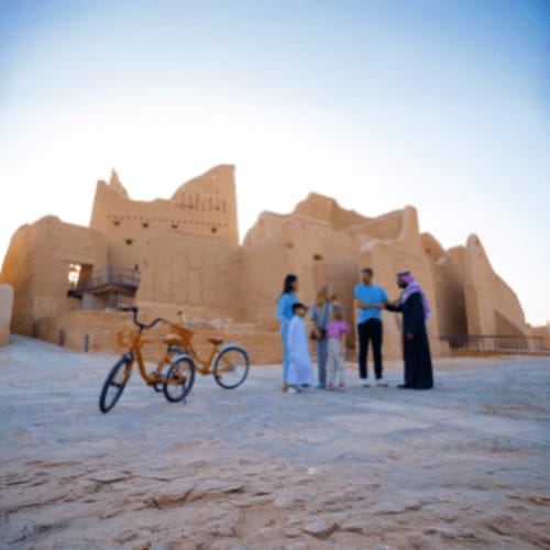 Tourists standing near a bicycle in front of an ancient mud brick fortress under clear blue sky in a desert setting