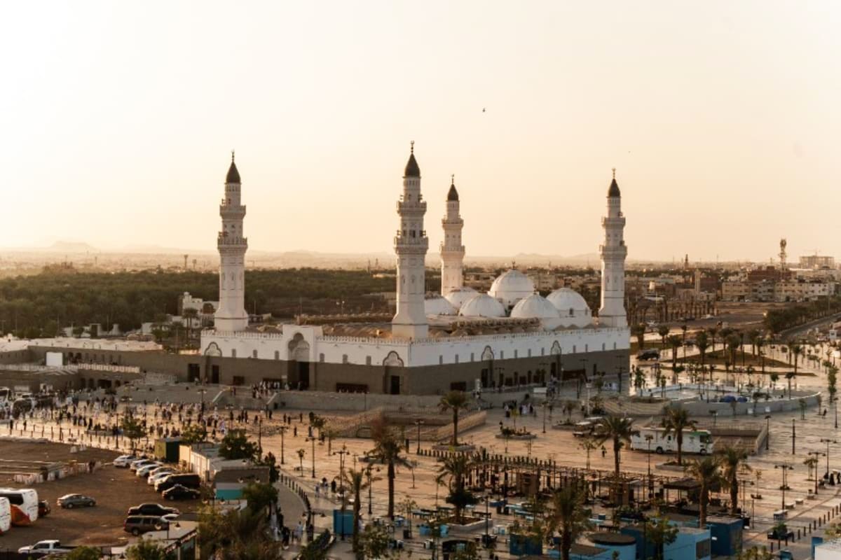 Quba Mosque in Medina with white domes and four minarets surrounded by palm trees and plaza with visitors