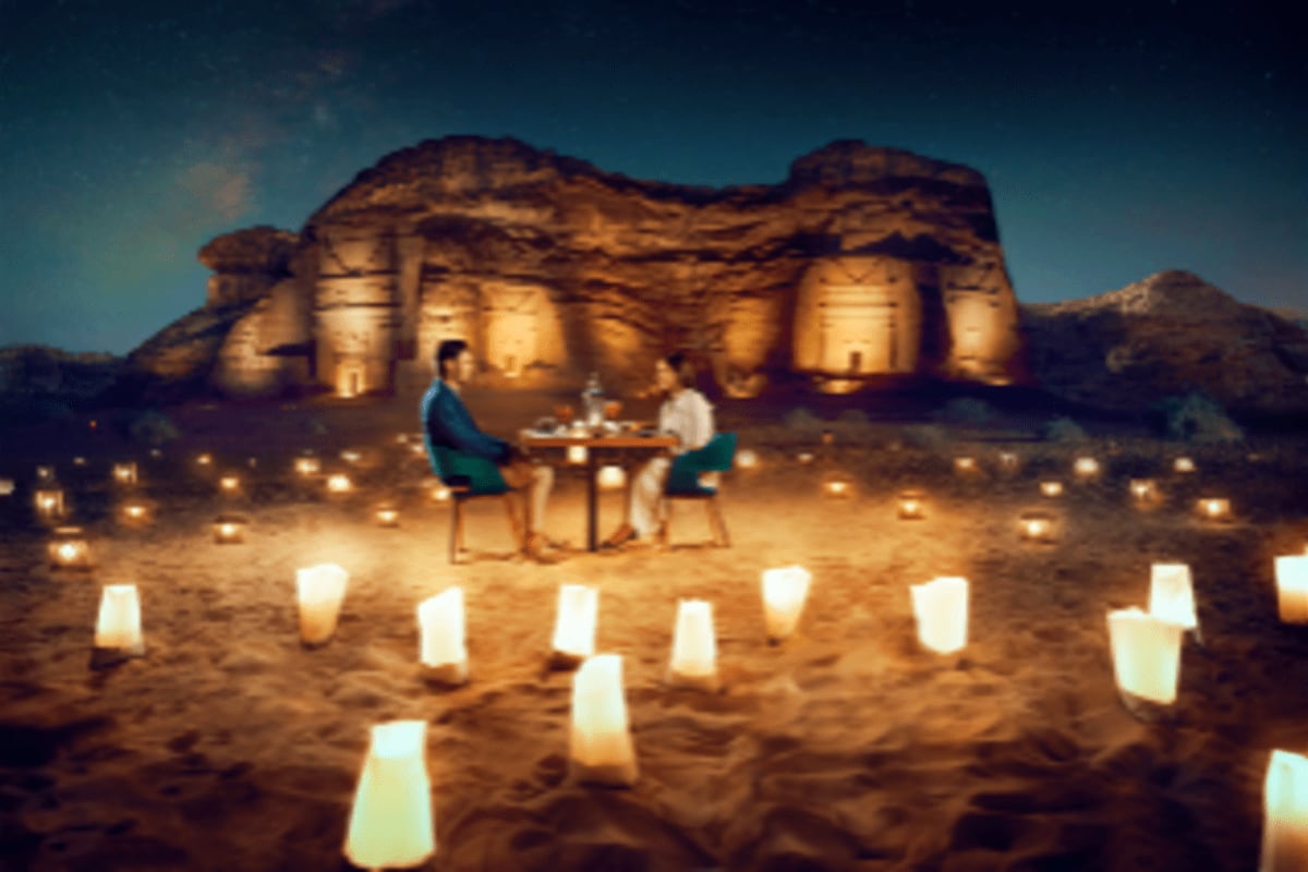 Couple dining at a table in the desert at night with lit candles surrounding them and an illuminated ancient stone structure