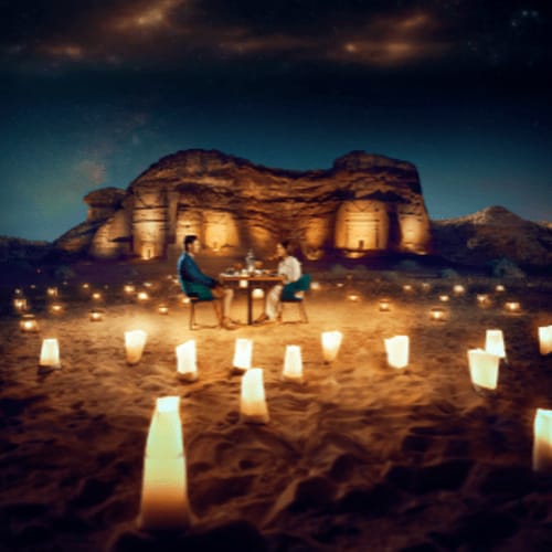 Couple dining at candlelit table in desert with illuminated sandstone rock formations at night in Saudi Arabia